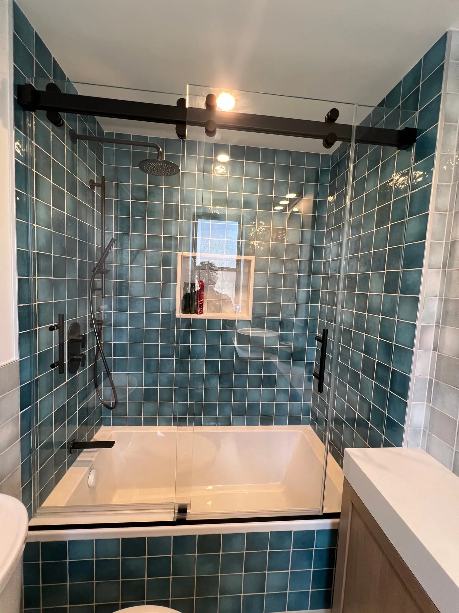 A deep green tiled tub and shower with a black barn-style sliding glass door and a wood vanity alongside