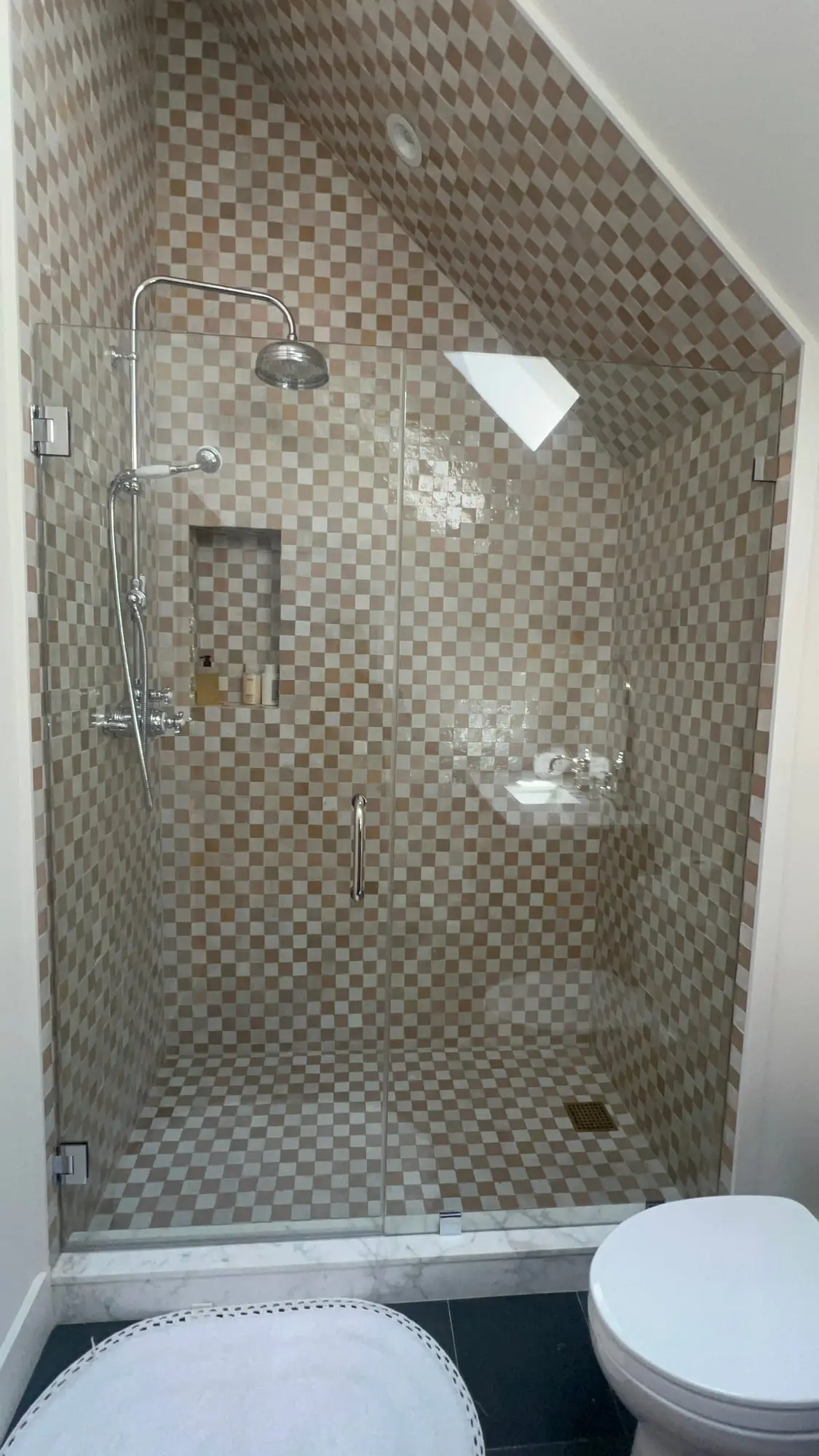 A shower fitted beneath a sloping ceiling, its glass panel cut to the angle of the roof, in small mosaic tile