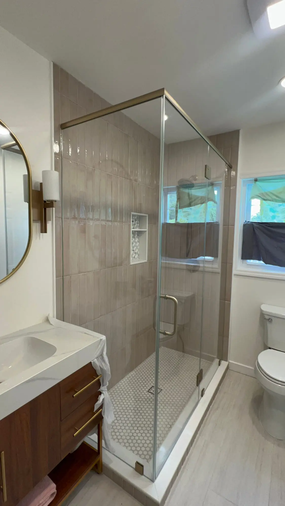 A frameless corner shower beside a window, with a round mirror above a wood vanity