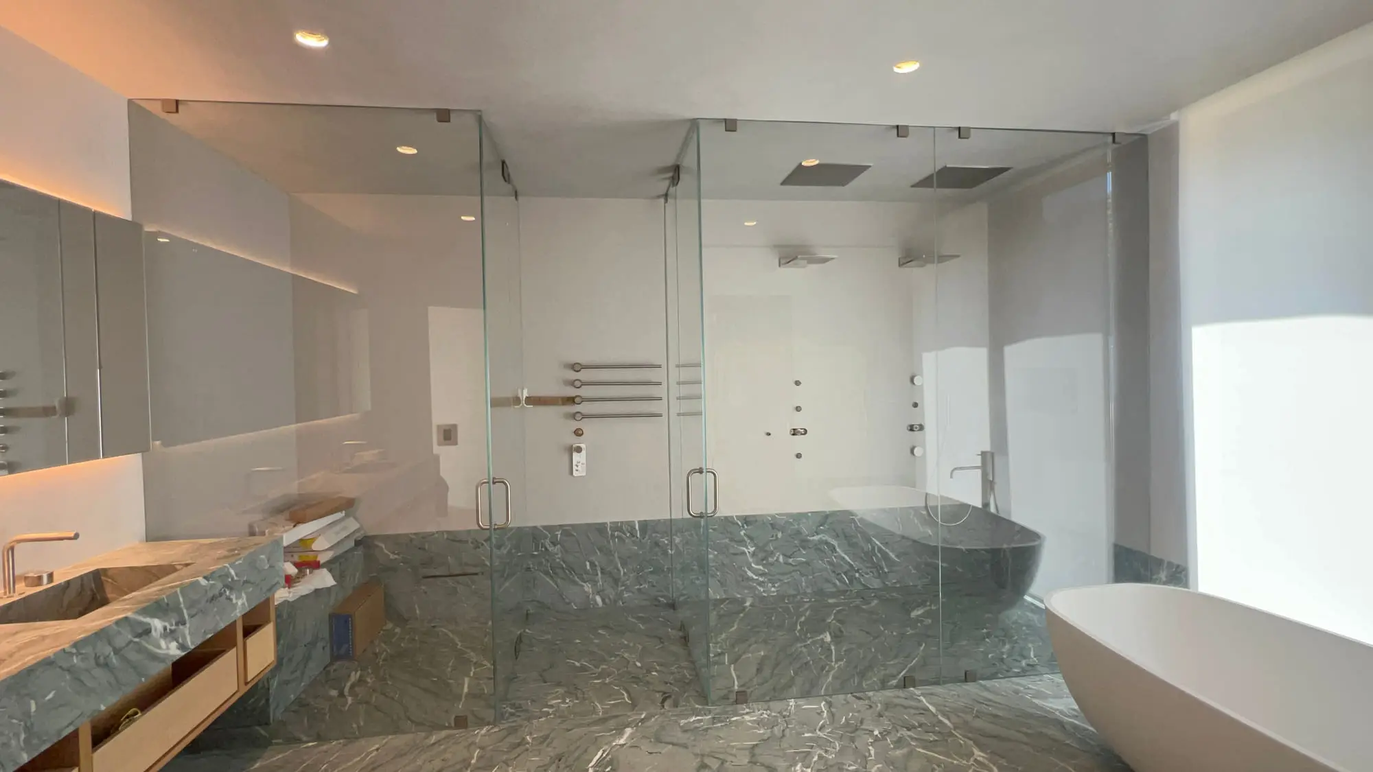 A wide bathroom with a green marble tub surround and a frameless glass panel separating the shower area