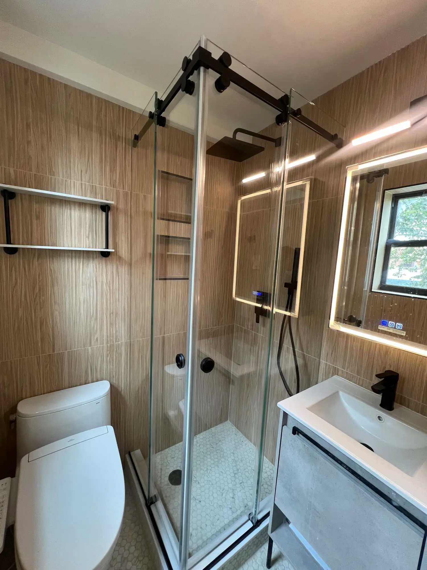 A corner shower with black framed sliding glass doors against a wood-panelled wall, with black towel rails