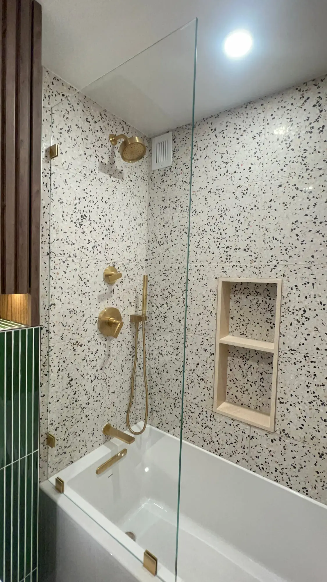 A bath with a fixed glass screen against a terrazzo tiled wall, with gold fittings and a fluted green accent panel