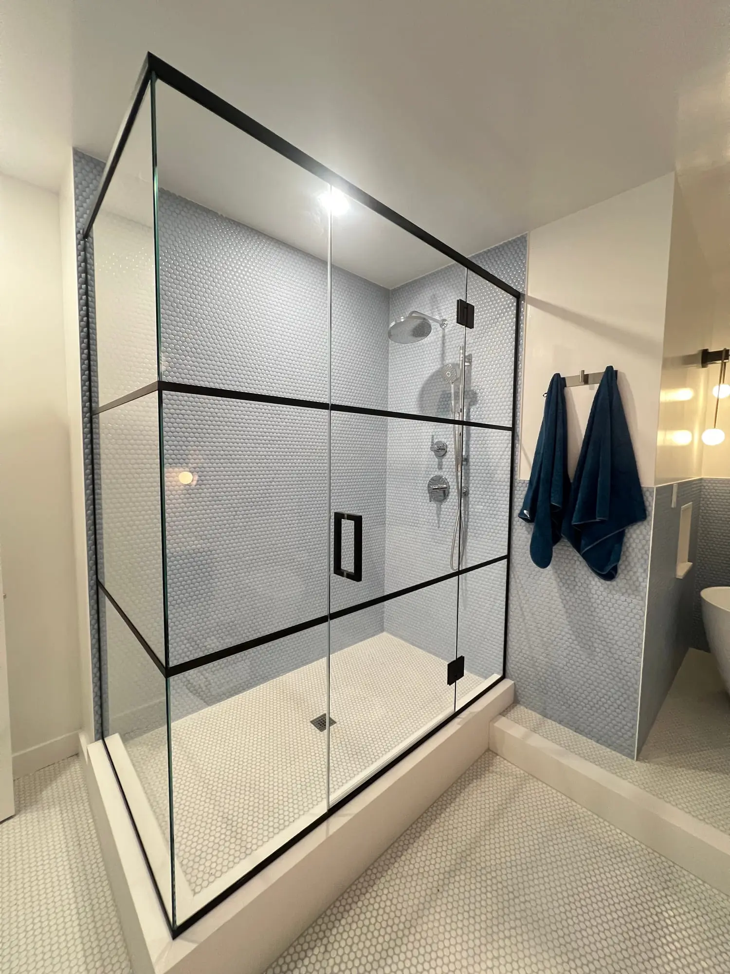 A large walk-in shower enclosed in clear glass divided by a black grid, in a pale marble bathroom with navy towels on a rail