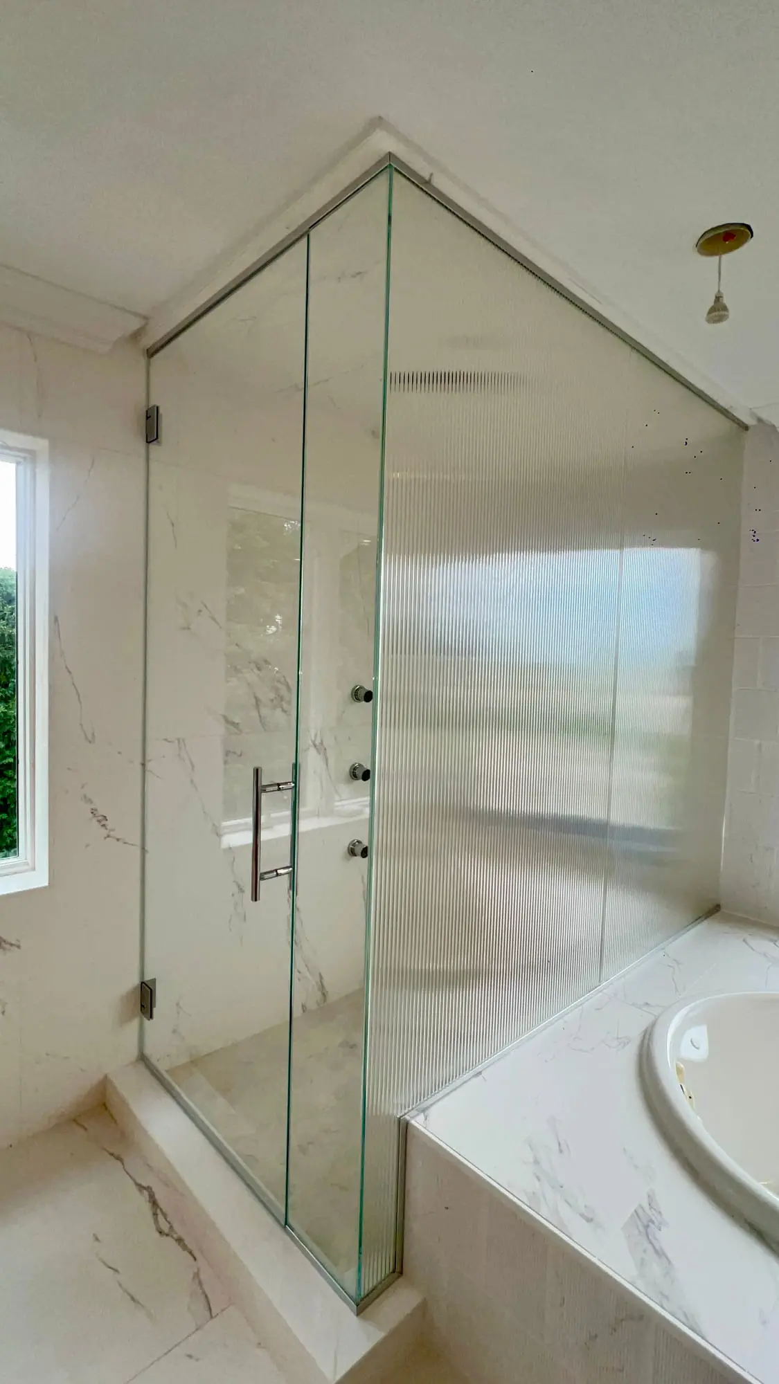 A glass corner shower next to a freestanding soaking tub, with gold fittings and a window between them