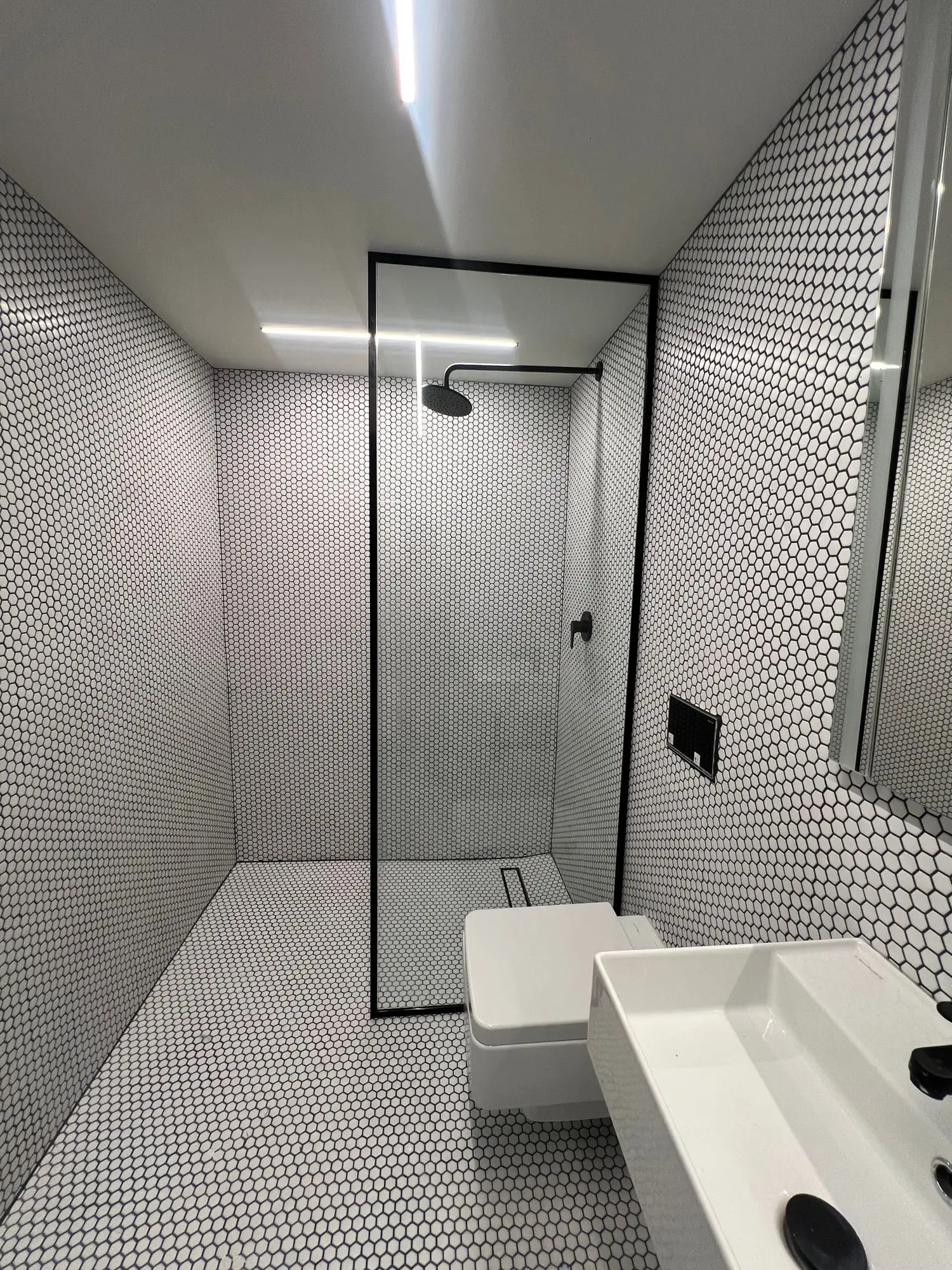 A walk-in shower in white penny-round tile framed in black, beside a wall-hung basin
