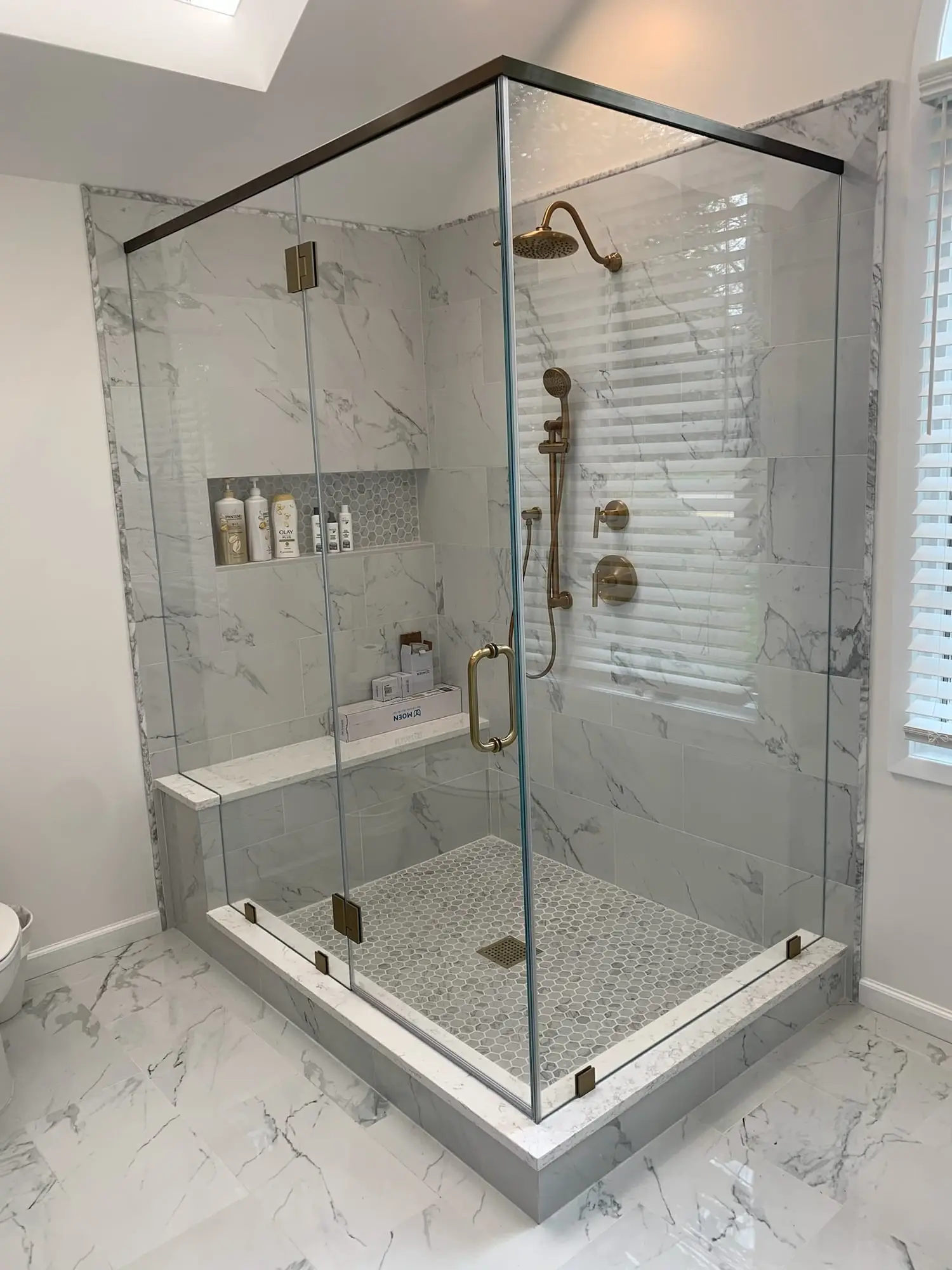 A frameless corner shower with brushed brass hinges, handle and header, a tiled bench and a pale marble floor