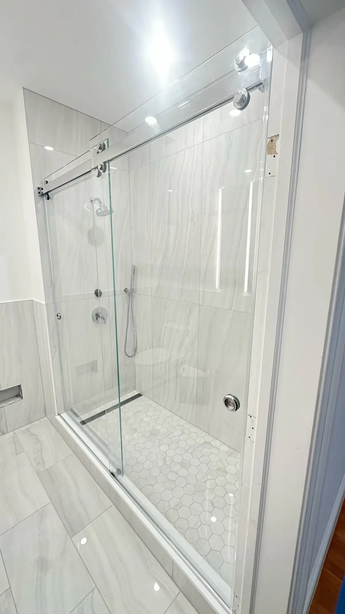 A frameless sliding glass door on a polished chrome roller track, across a shower in large white porcelain tile