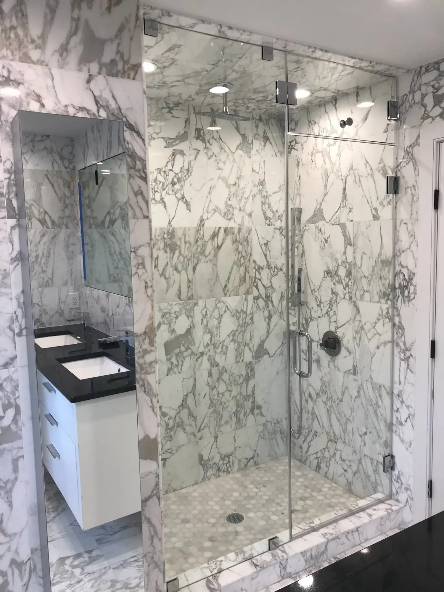 A tall frameless door and panels enclosing a walk-in shower lined in boldly veined white marble, beside a floating vanity