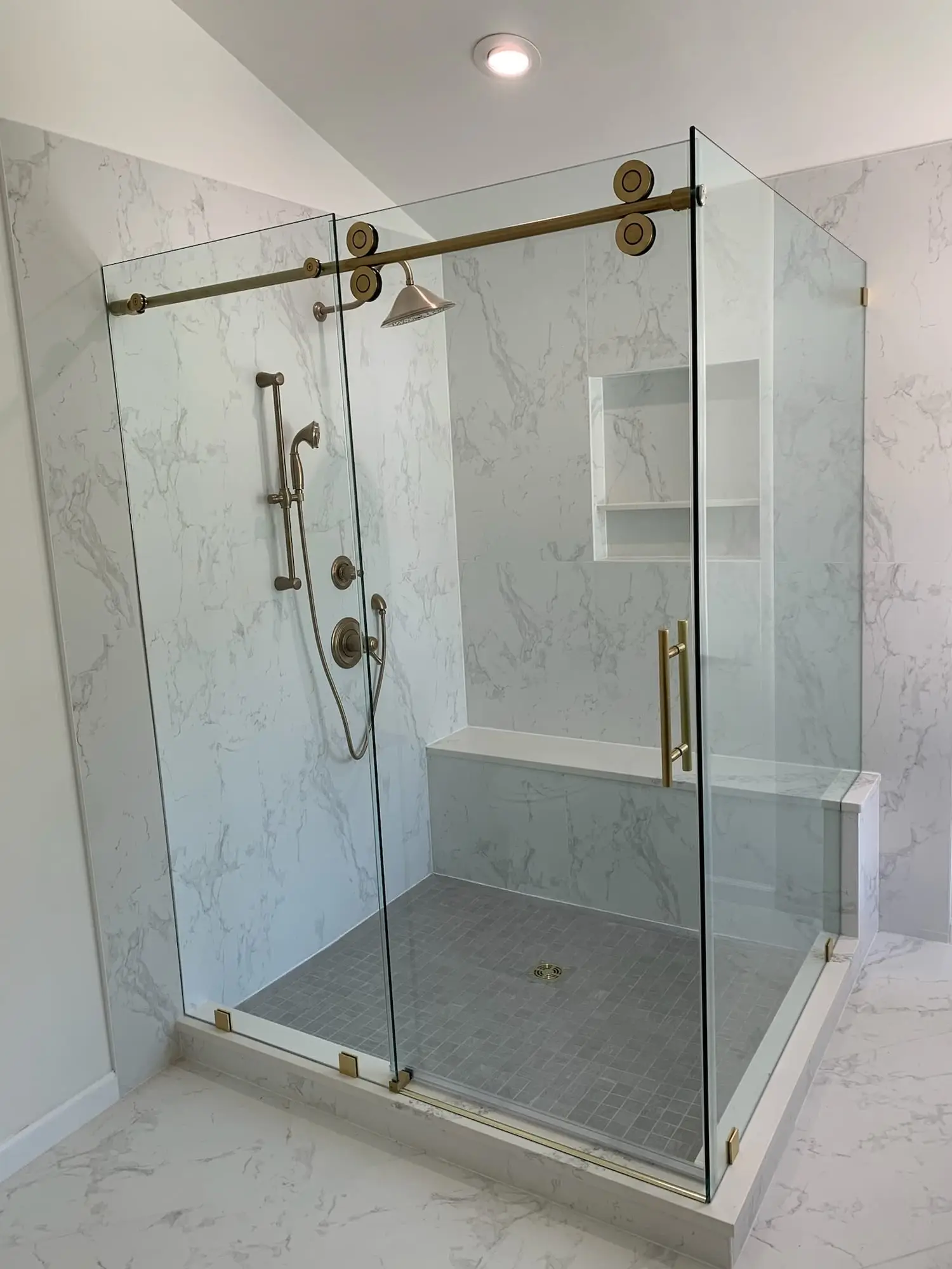 A corner shower with a sliding glass door on exposed brass rollers, a brass handle and a built-in bench