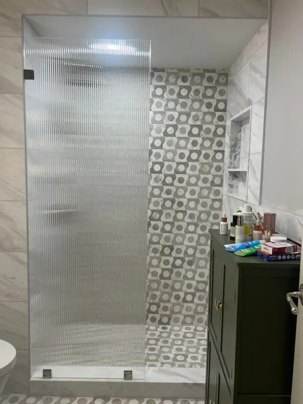 A single fluted glass panel screening a walk-in shower, against a wall of patterned grey and white tile