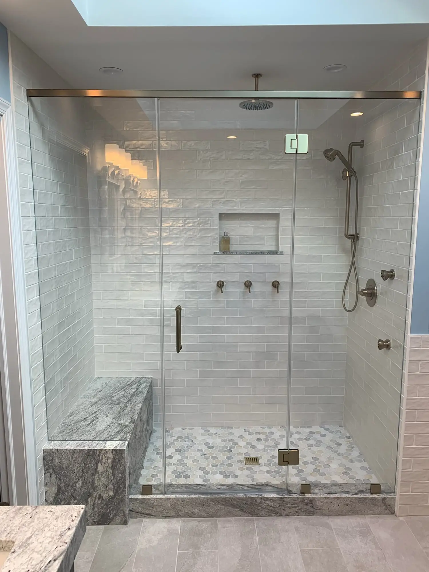 A wide frameless enclosure with a door, fixed panels and a satin nickel header, in front of a marble bench and a pebble floor
