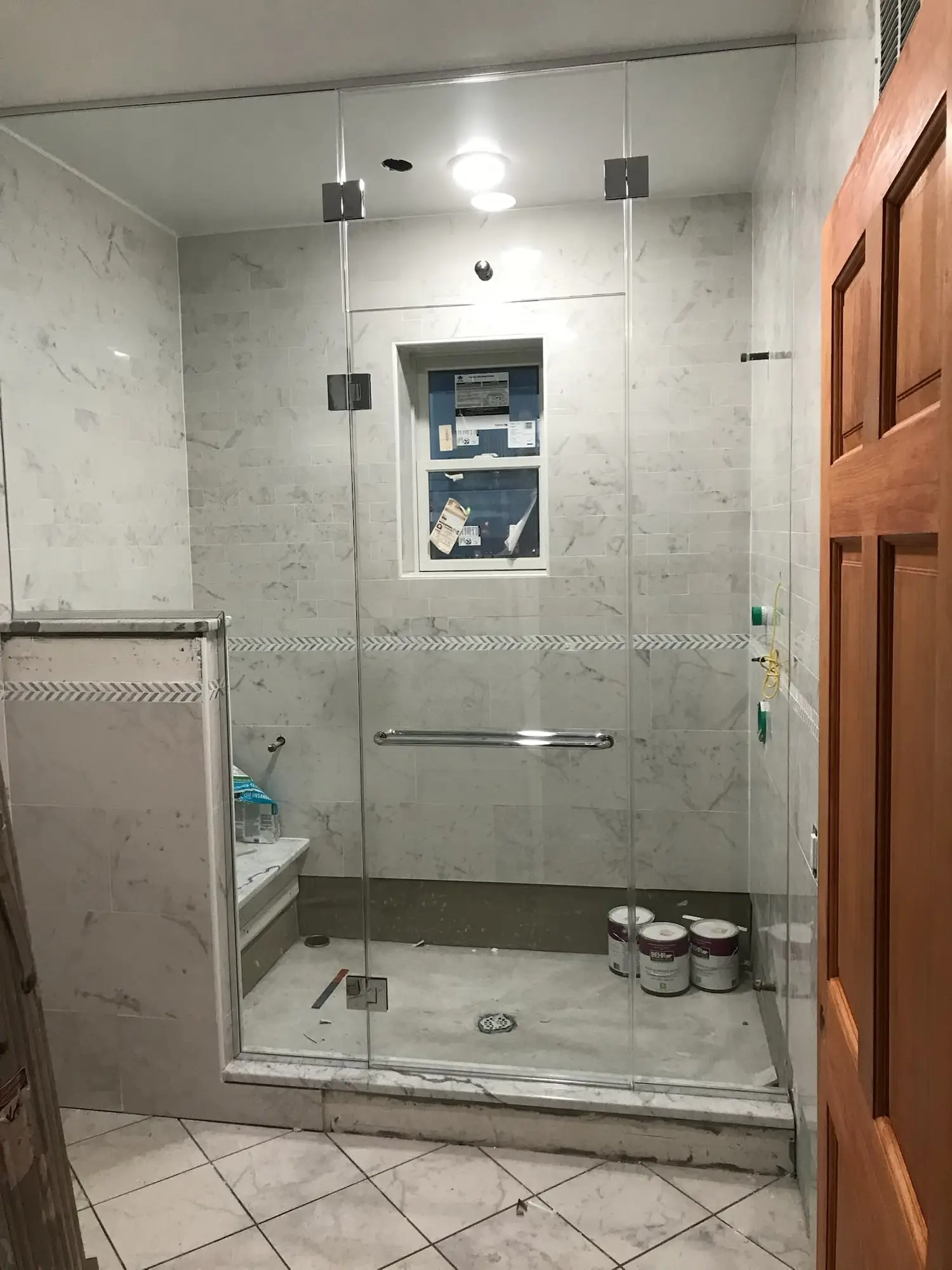 A frameless door and fixed panel with a towel bar on the glass, enclosing a marble shower with a lit niche