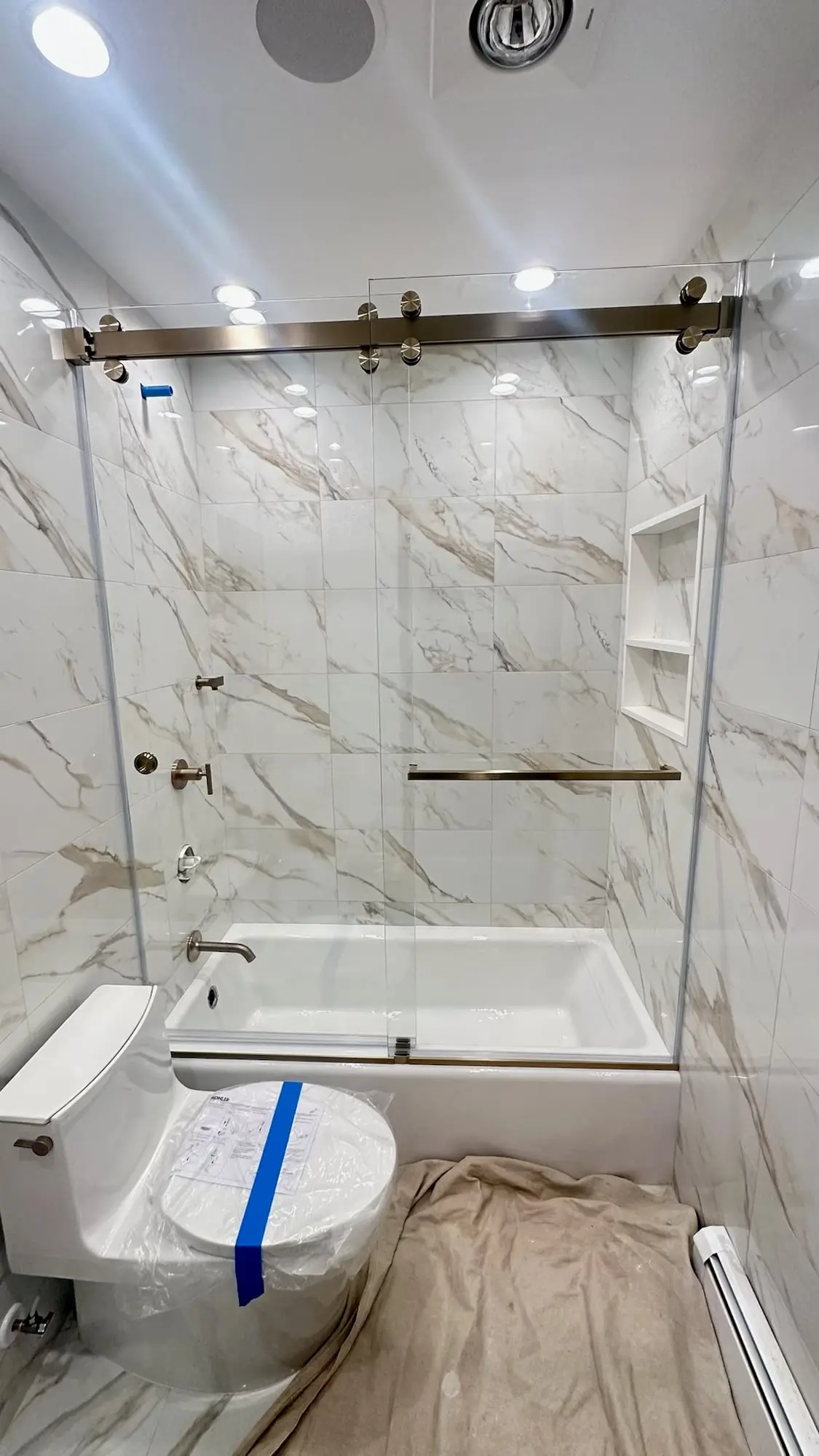 A sliding glass door on brushed gold rollers across a soaking tub, in a white marble bathroom