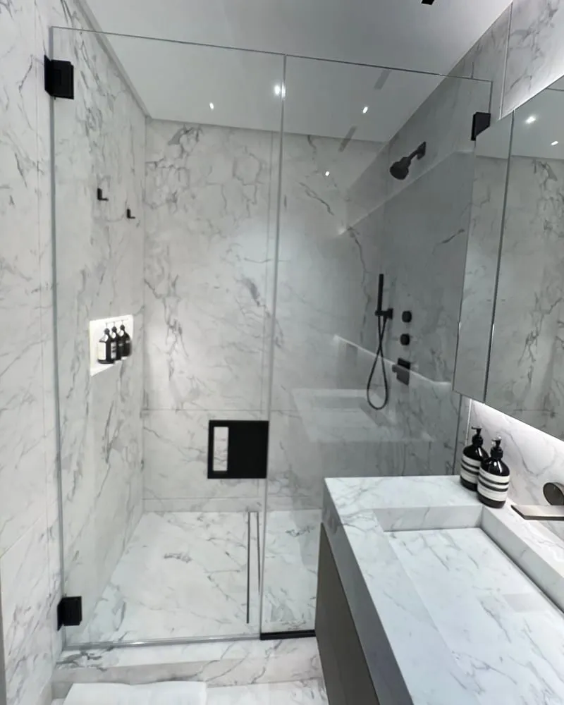 A frameless glass shower lined in white marble with black fixtures, beside a marble vanity